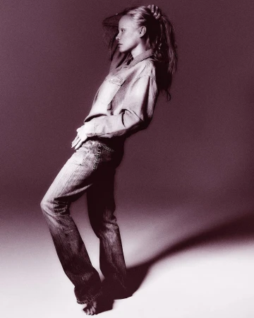 Blonde woman in denim shirt and jeans poses barefoot against a solid backdrop in a studio setting with soft lighting, fashion portrait.
