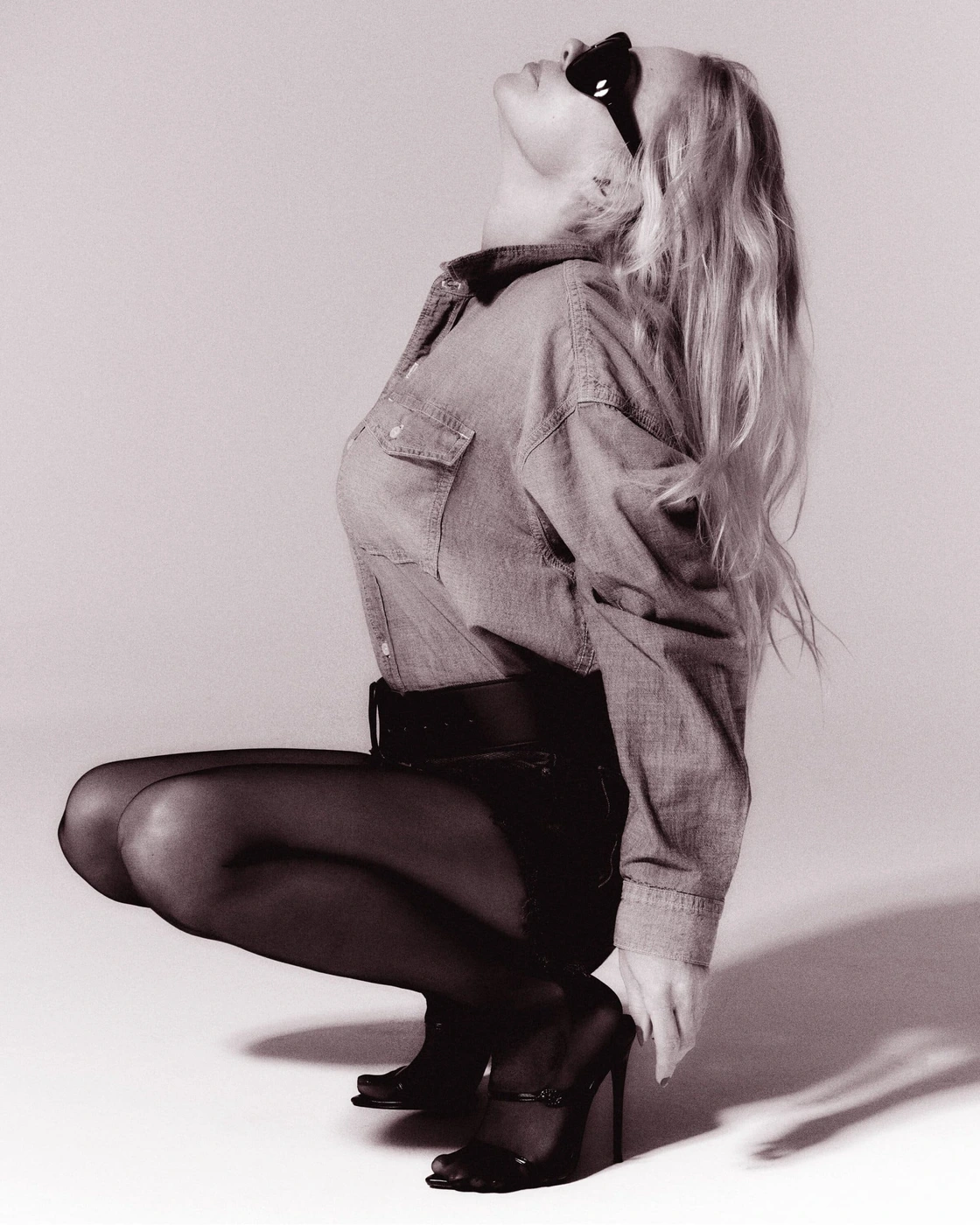 Model in denim shirt and dark stockings crouching against a neutral background wearing sunglasses.