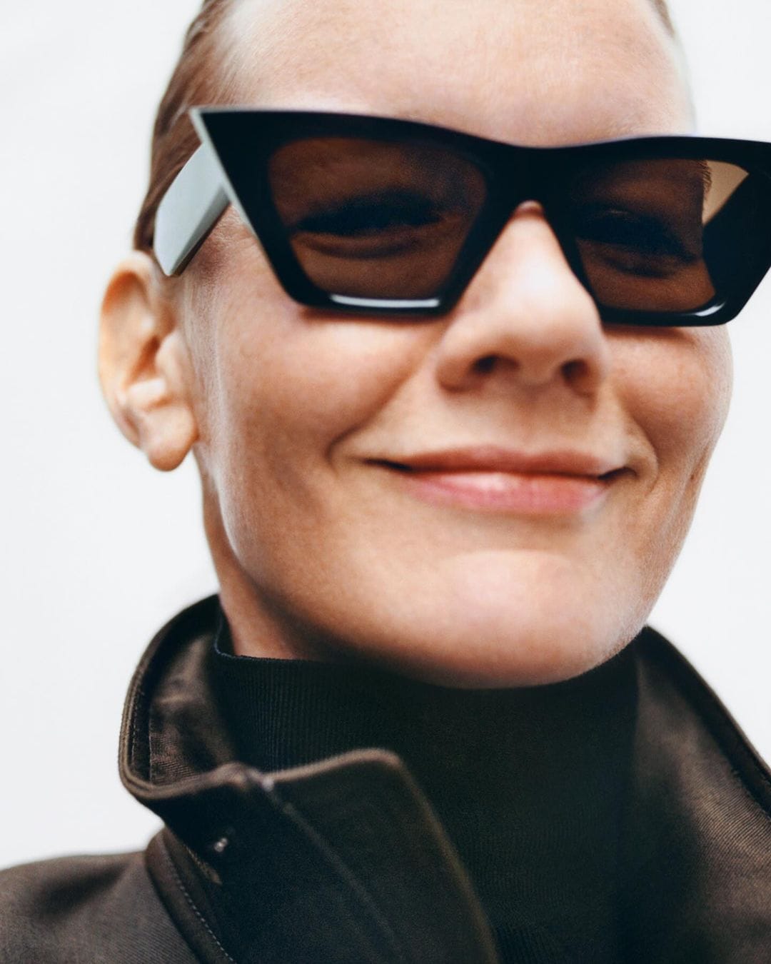 Close-up portrait of an adult female smiling and wearing black sunglasses and a dark brown jacket against a white background.