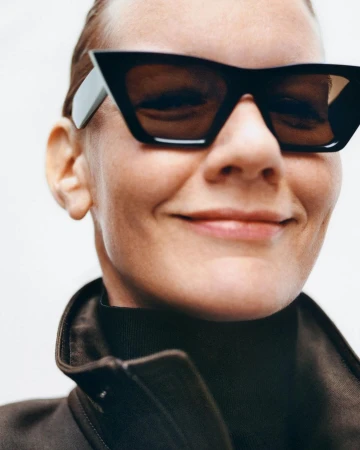 Close-up portrait of a woman wearing angular black sunglasses and a dark high-neck jacket against a neutral backdrop.