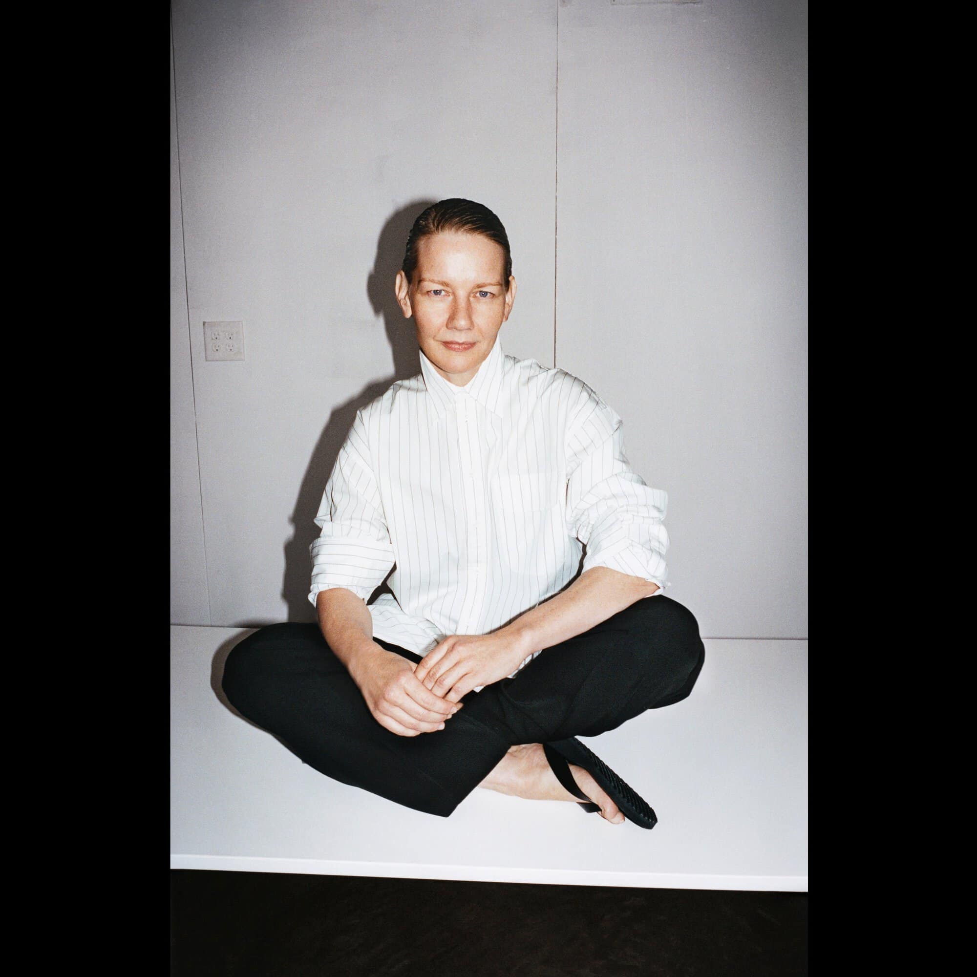 Portrait of a slim adult with bare feet sitting cross-legged on a platform, wearing a striped shirt and dark pants against a neutral backdrop.