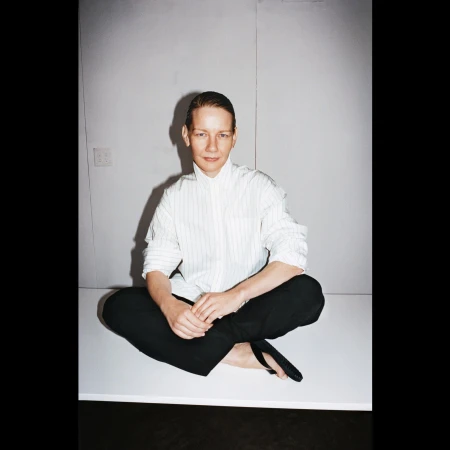 Portrait of a slim adult with bare feet sitting cross-legged on a platform, wearing a striped shirt and dark pants against a neutral backdrop.