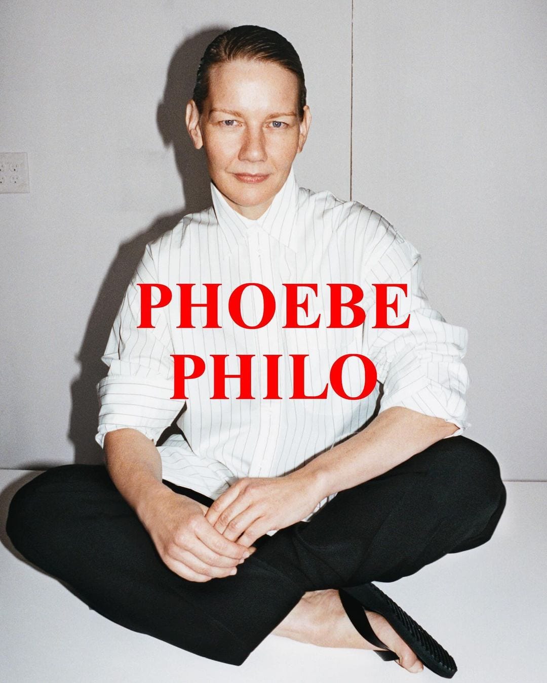 A fair-skinned person with short hair sits cross-legged on a white surface wearing a striped shirt and black pants, facing the camera.
