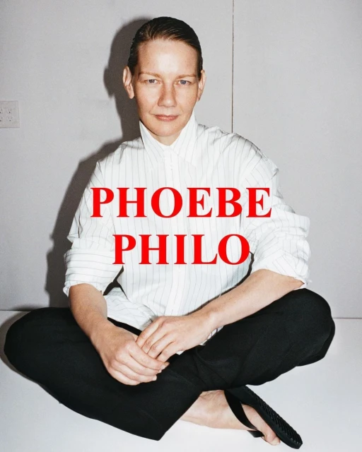 A fair-skinned person with short hair sits cross-legged on a white surface wearing a striped shirt and black pants, facing the camera.