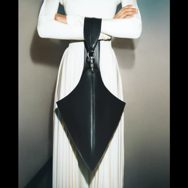 Fashion shoot featuring a model in a white dress showcasing a black, geometric leather handbag with metallic ring detail and unique shape.
