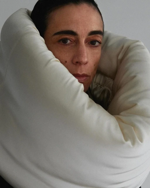 Headshot of a woman with fair skin, dark hair, and neutral expression wrapped in a cream-colored padded garment against a plain background.