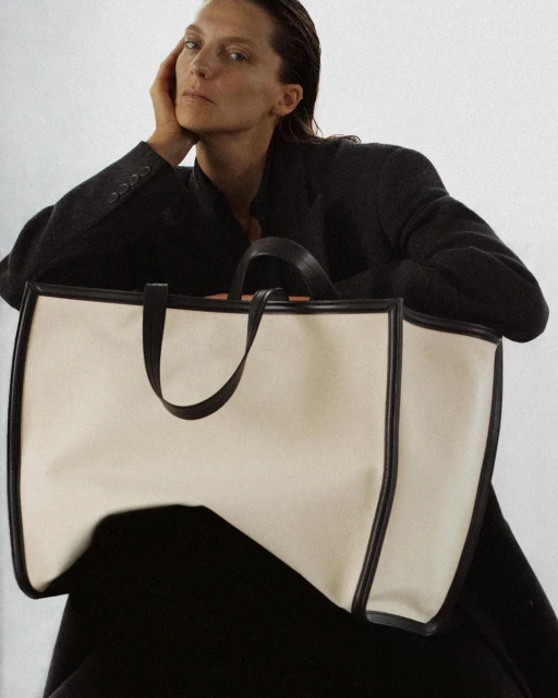 Woman poses with a large tote bag in a studio setting. The bag is cream with black trim, and the woman wears a dark blazer.