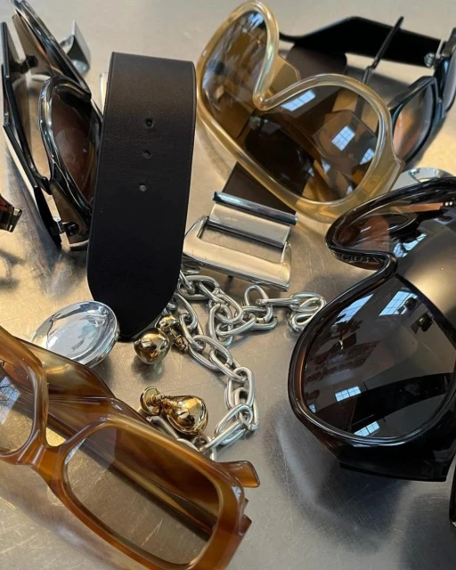 Still life of various pairs of sunglasses, a black belt, and a silver chain on a metallic surface, showcasing fashion accessories.