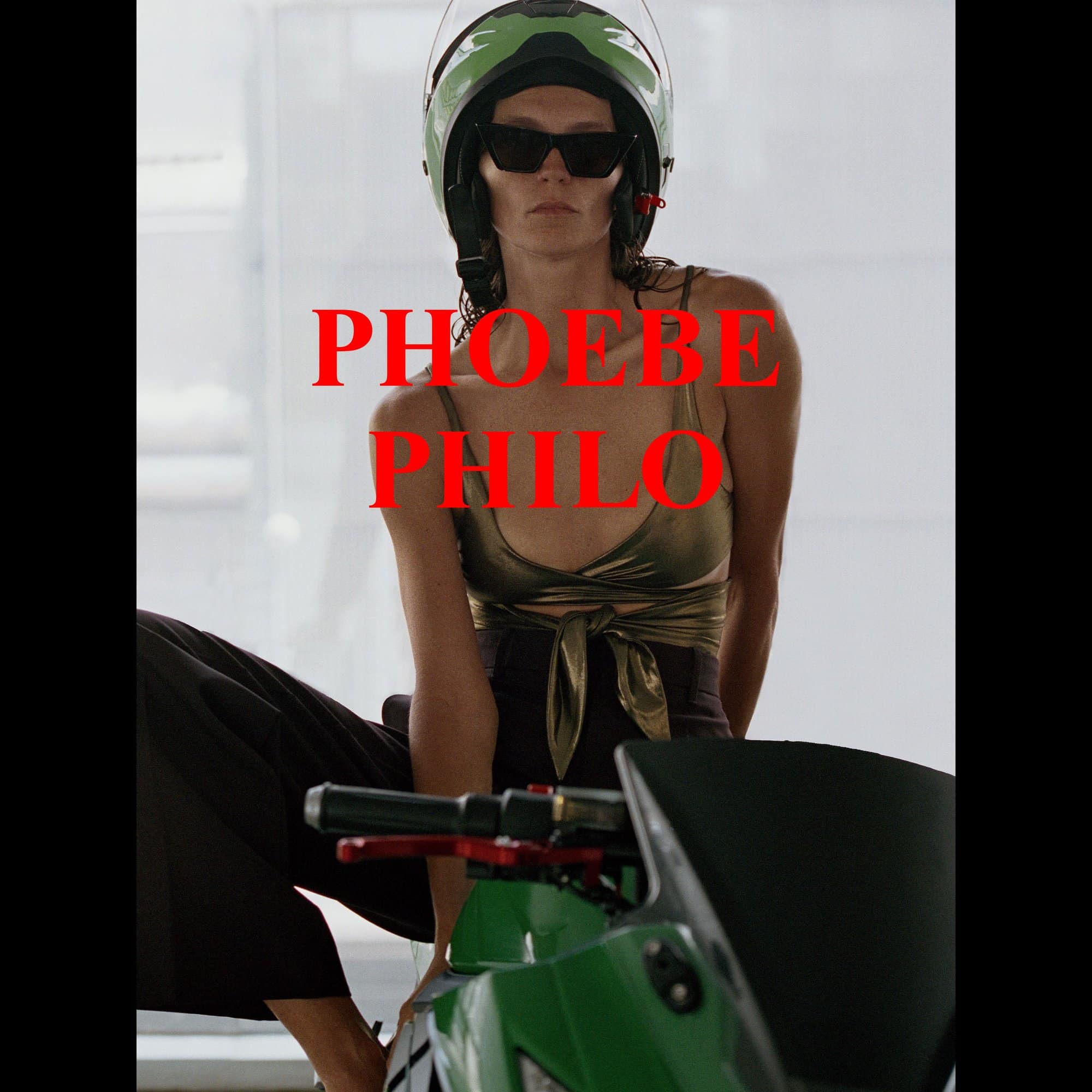 A fashion shot of an adult woman in a spaghetti-strap top and helmet, sitting on a green motorcycle, wearing black sunglasses.