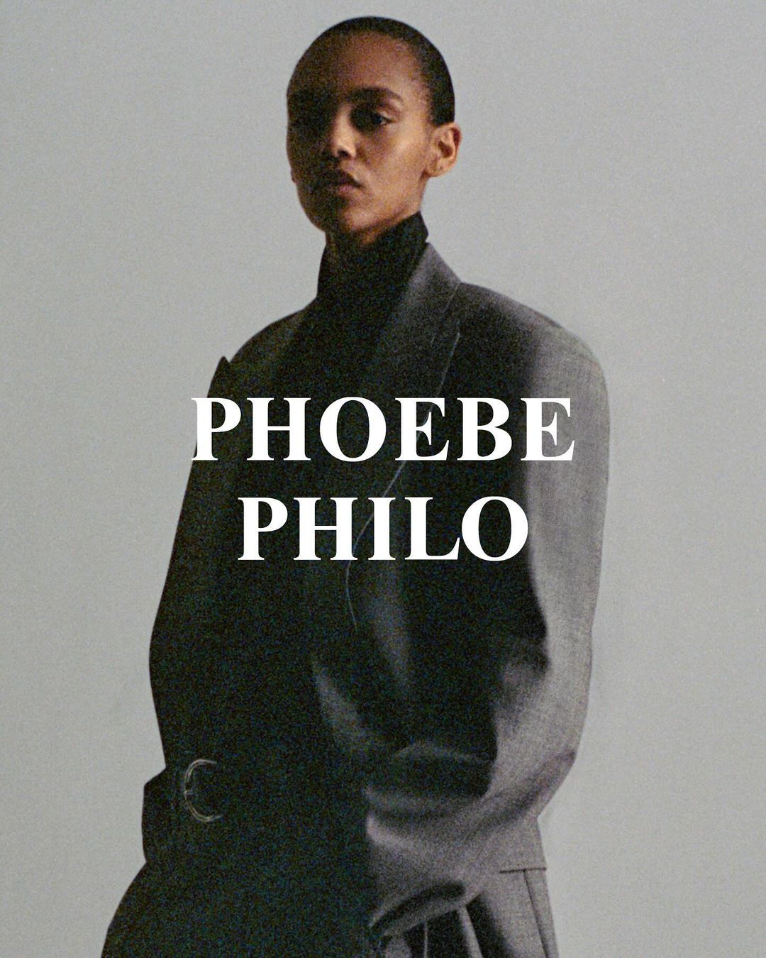 Fashion portrait of a woman wearing a grey jacket and black turtleneck against a neutral background with the text PHOEBE PHILO.