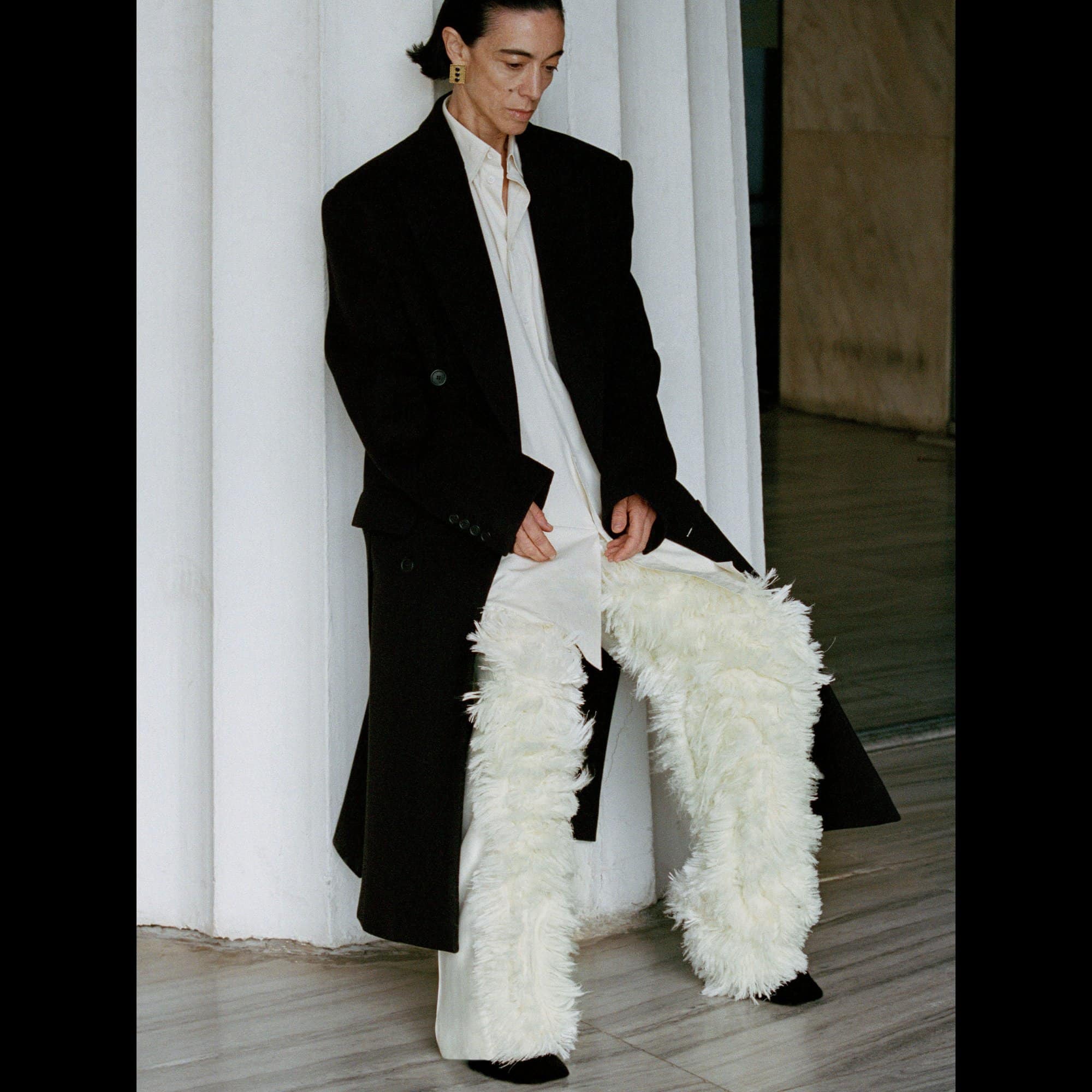 Fashionable adult poses in a black coat and white faux-fur pants in front of a modern concrete wall in an editorial fashion photograph.