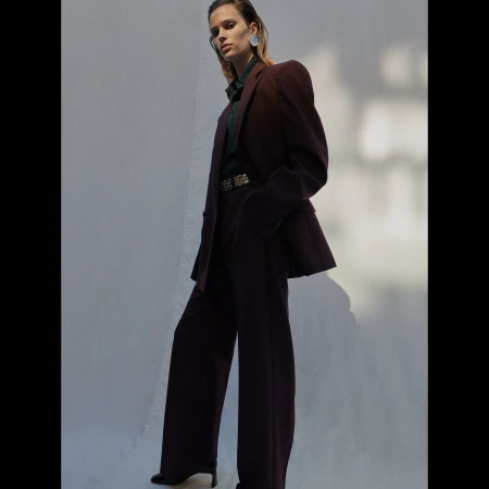 Model wearing a dark burgundy suit and statement earrings standing against a white backdrop.