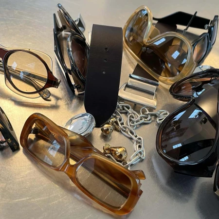Arrangement of designer sunglasses, a black leather belt, and silver chain accessories on a metallic surface