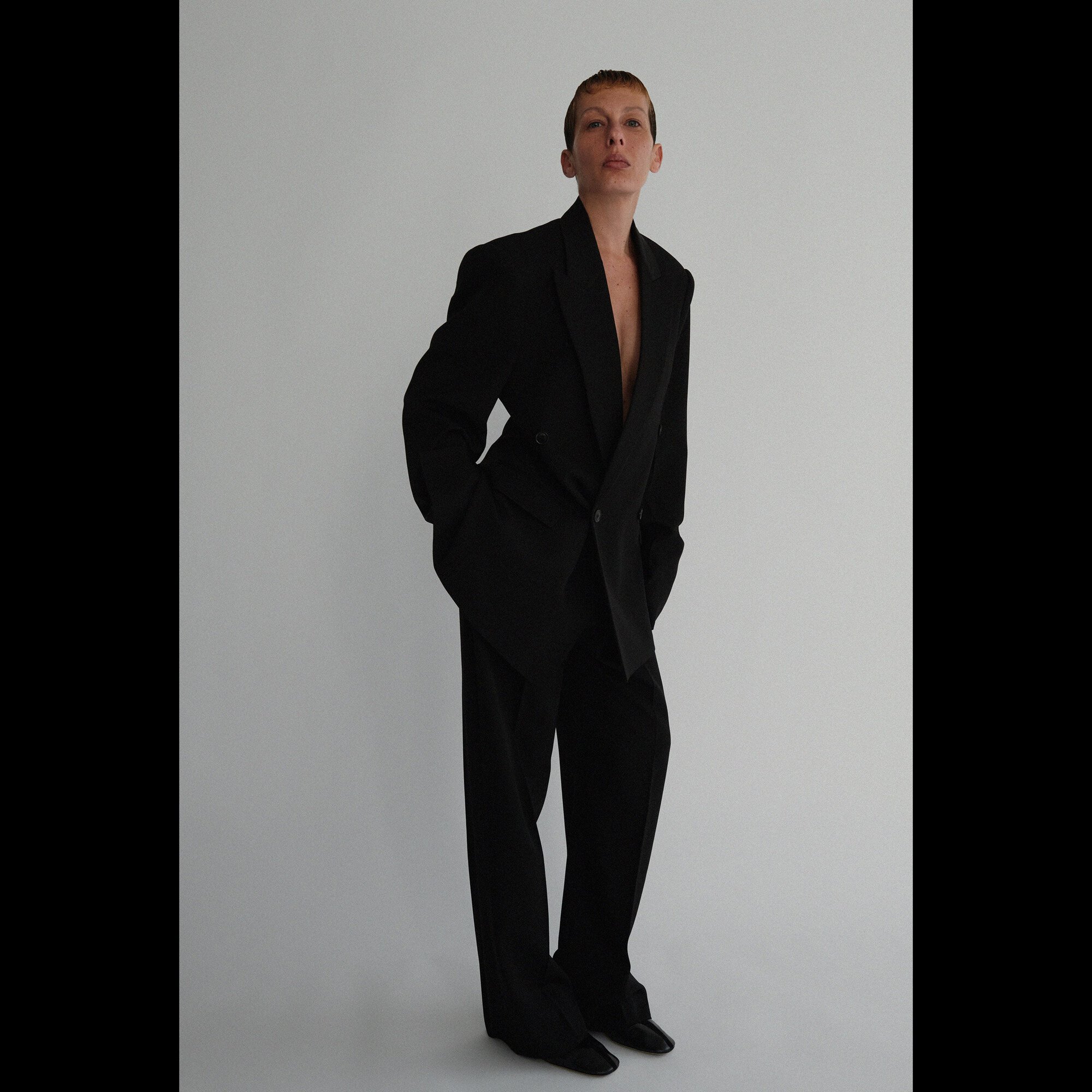 Full-length shot of a model in a black oversized suit posing in a studio with a neutral background, looking directly at the camera.