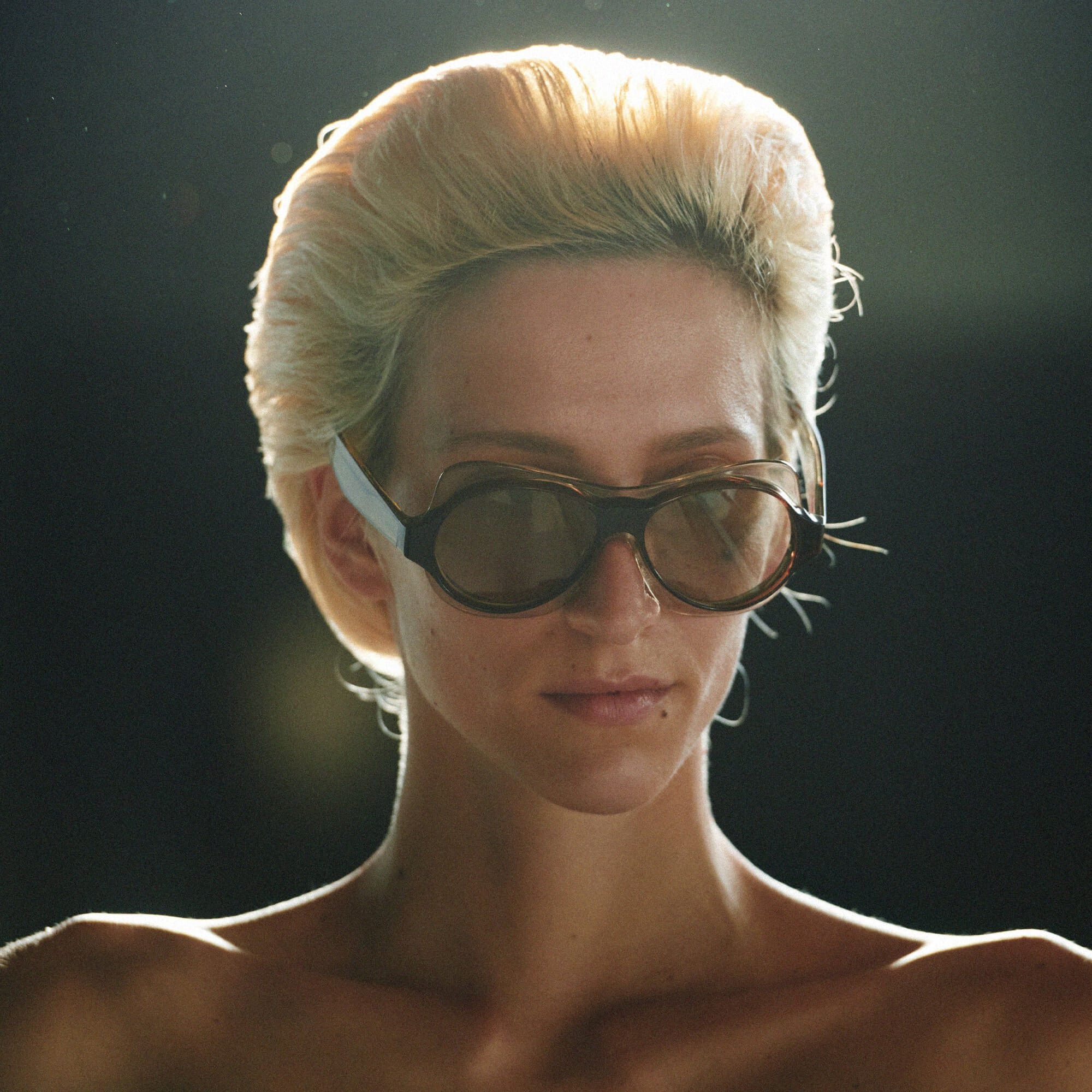 Close-up portrait of a woman with short blonde hair and unique eyewear against a dark background with soft, directional lighting.