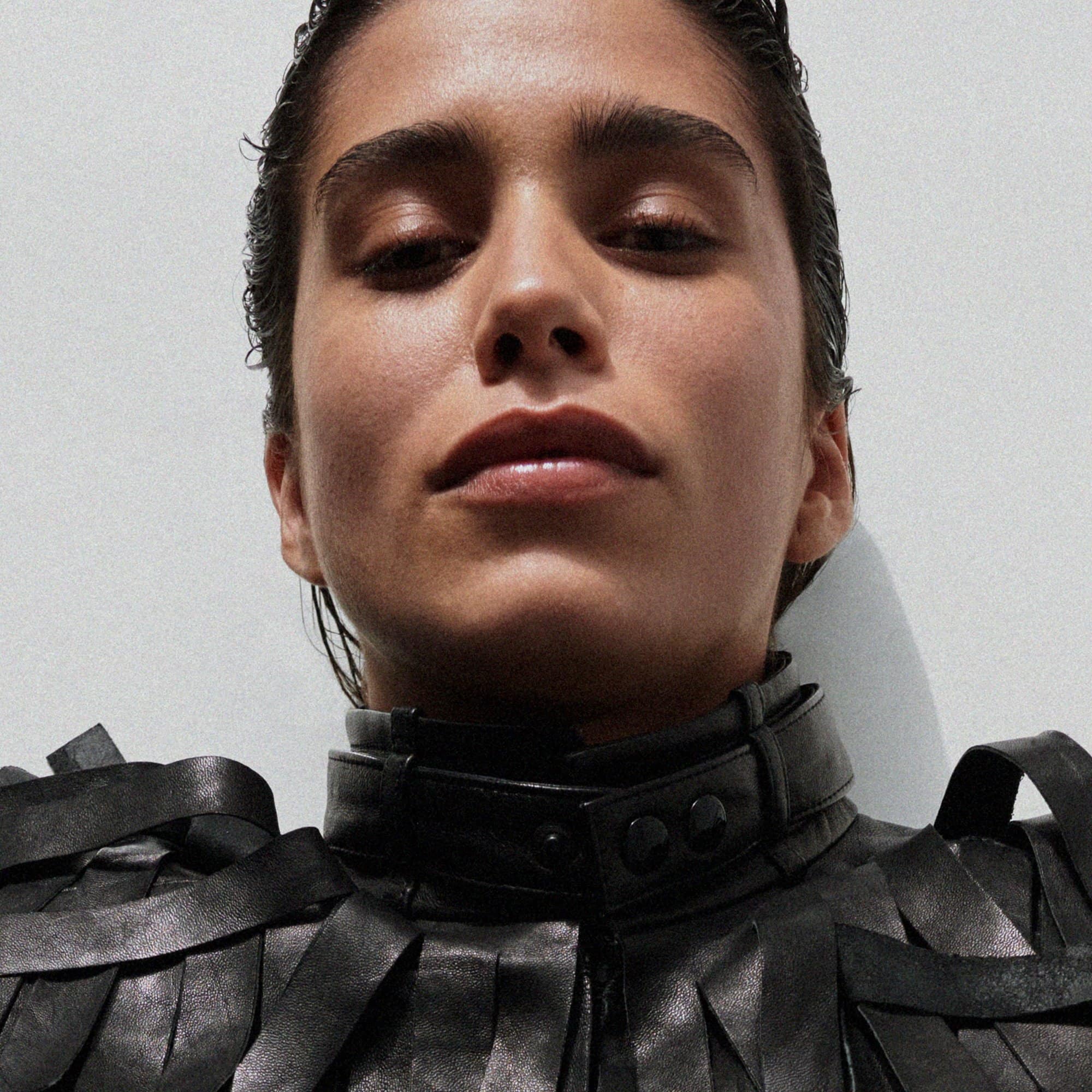 Close-up portrait of a woman with wet-look hair in a black leather jacket with unique collar against a plain light background.