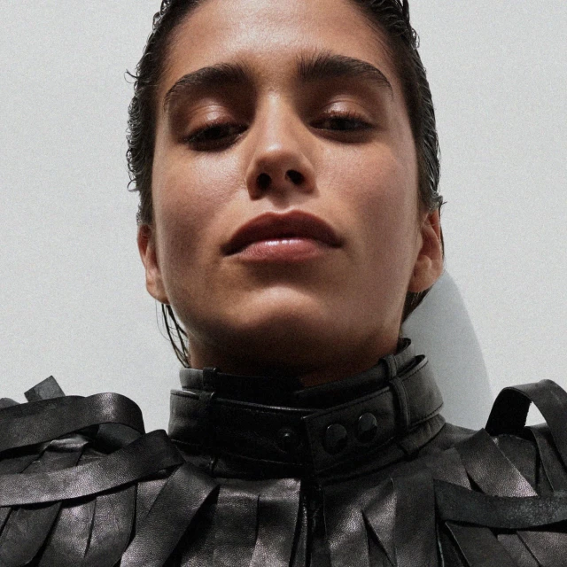 Close-up portrait of a woman with wet-look hair in a black leather jacket with unique collar against a plain light background.