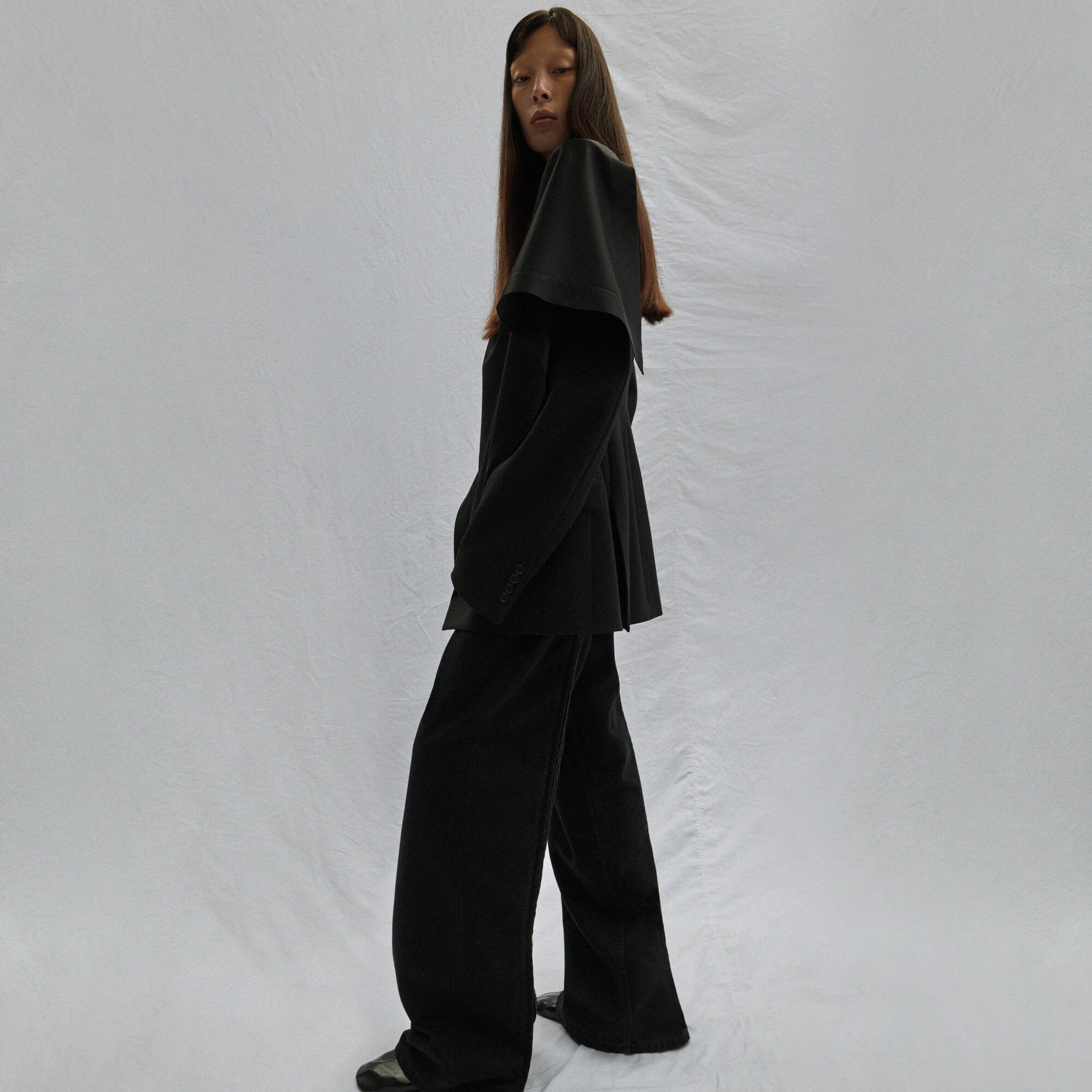 Full-length fashion shot of an Asian female model in a black suit with a cape-like collar against a neutral backdrop in a studio setting.