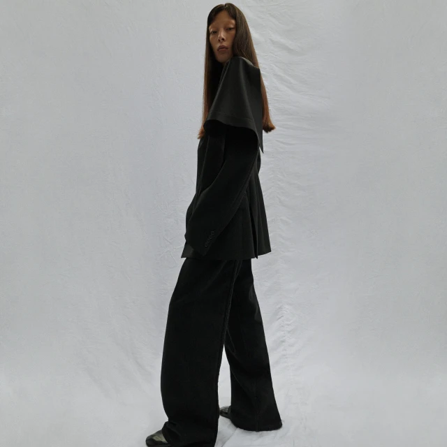 Full-length fashion shot of an Asian female model in a black suit with a cape-like collar against a neutral backdrop in a studio setting.