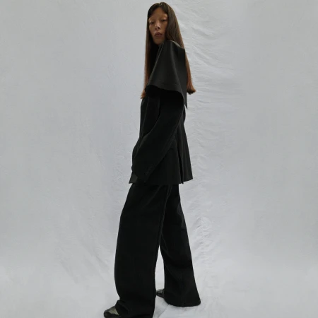 Full-length fashion shot of an Asian female model in a black suit with a cape-like collar against a neutral backdrop in a studio setting.