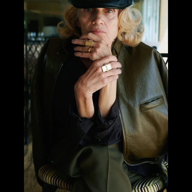 Stylish senior woman with olive leather jacket, black top, gold rings posing indoors, adding a touch of elegance to mature fashion.