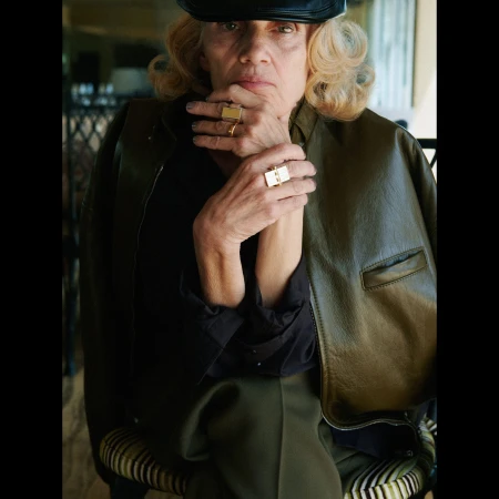 Stylish senior woman with olive leather jacket, black top, gold rings posing indoors, adding a touch of elegance to mature fashion.