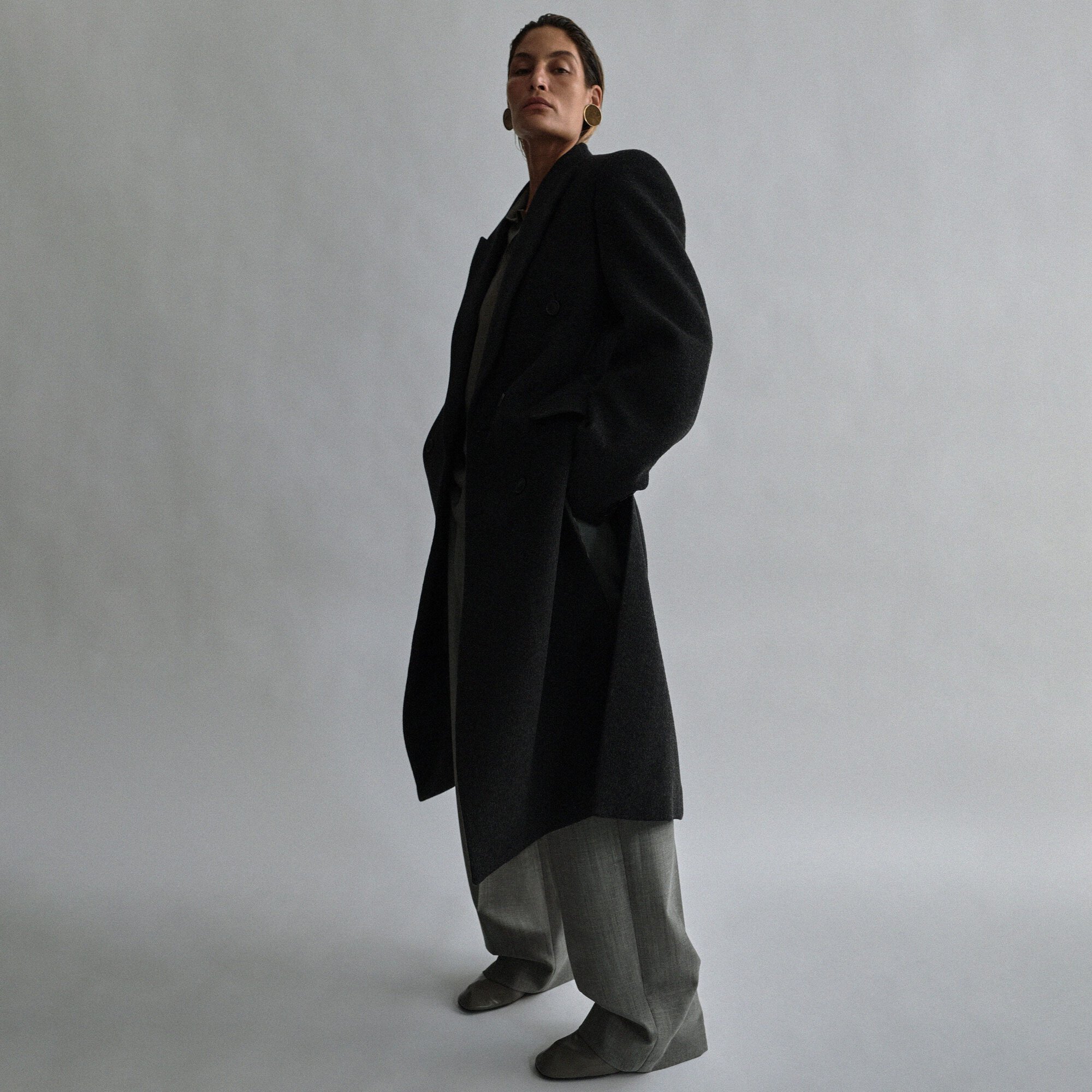 A woman in a black coat poses in a low-angle shot, featuring a neutral gray background and minimalist studio setting, showcasing fashion.