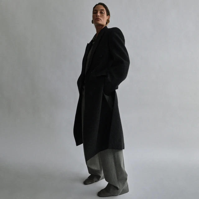 A woman in a black coat poses in a low-angle shot, featuring a neutral gray background and minimalist studio setting, showcasing fashion.