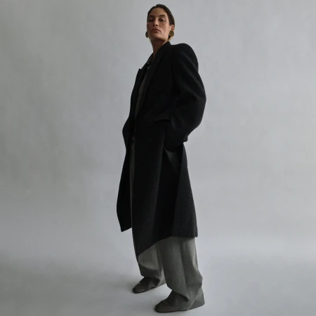 A woman in a black coat poses in a low-angle shot, featuring a neutral gray background and minimalist studio setting, showcasing fashion.