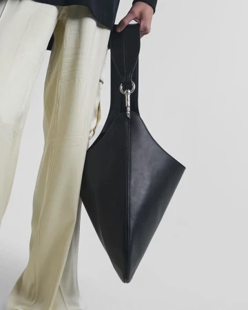 A close-up shot of a black leather handbag held by a person wearing cream trousers against a simple white backdrop.
