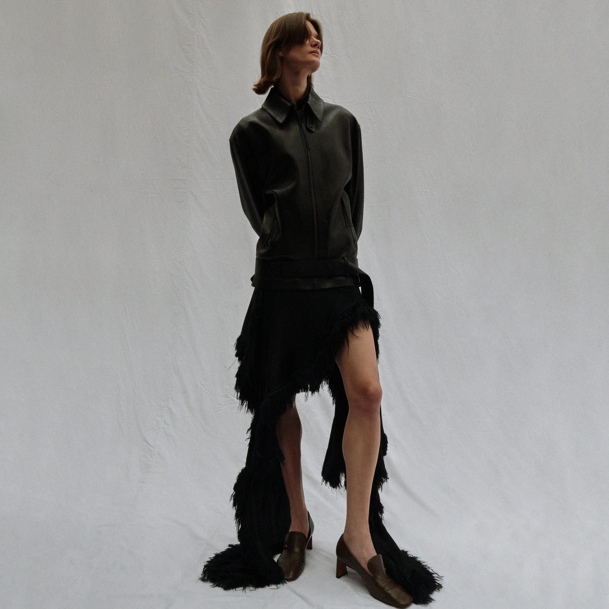 Adult posing in a photo studio wearing a dark jacket, black fringed skirt, and brown heeled loafers on a white background.