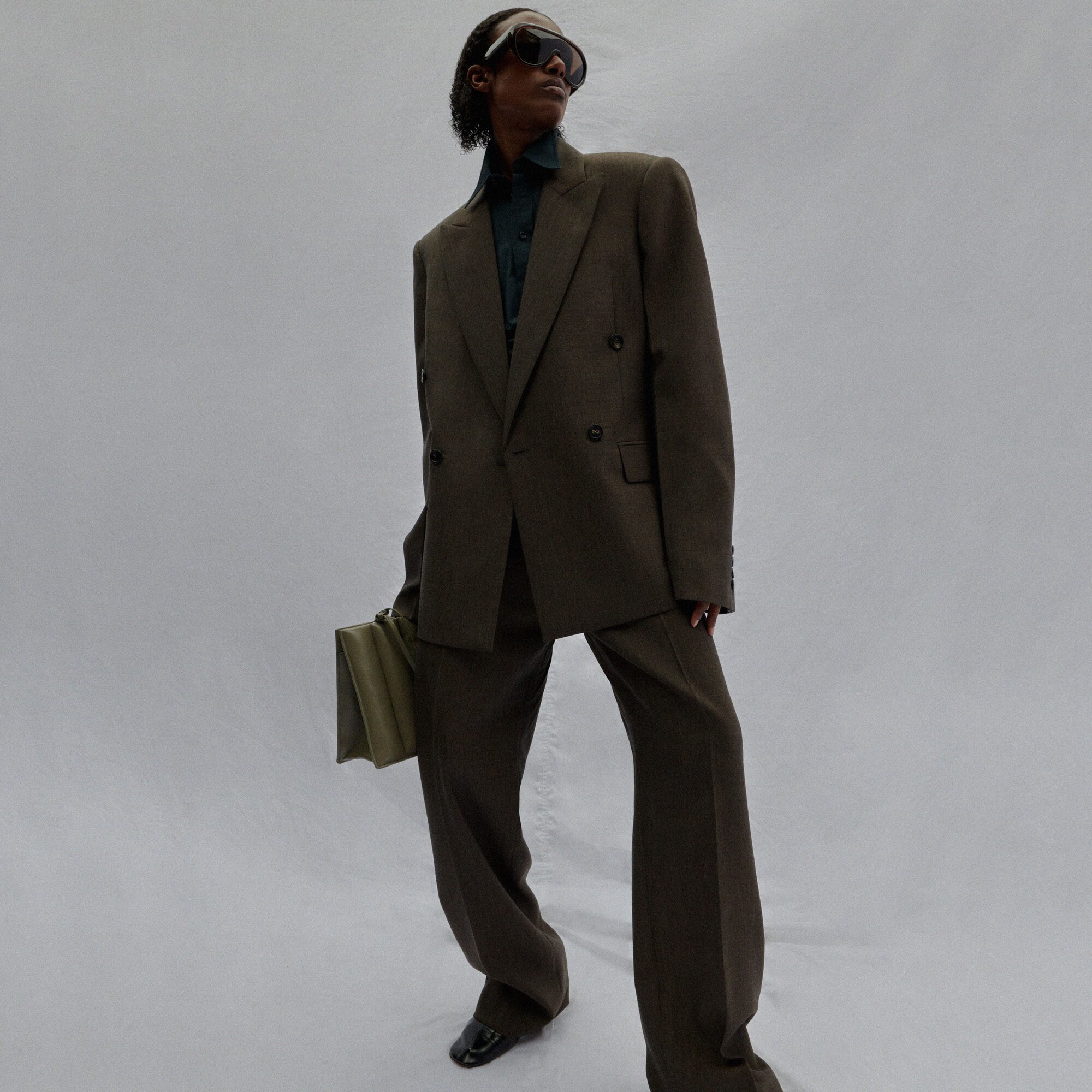 An adult male wearing a dark green suit and sunglasses is holding an olive green clutch in a studio with a white background.
