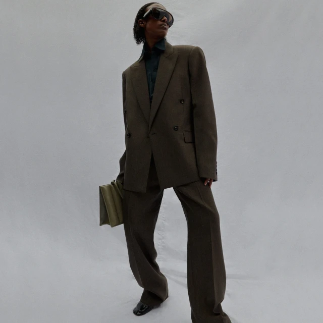 An adult male wearing a dark green suit and sunglasses is holding an olive green clutch in a studio with a white background.