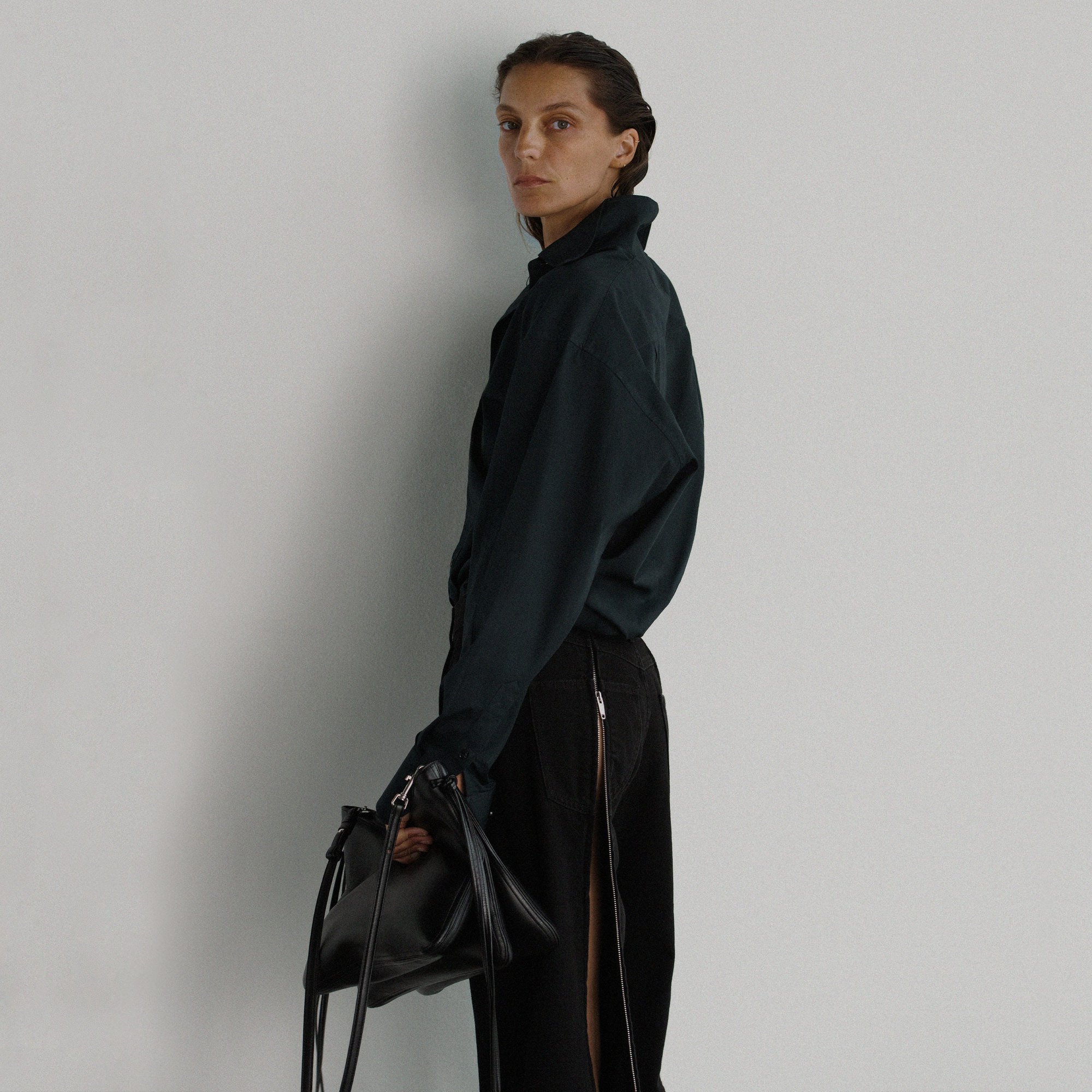 A woman in a black top and skirt holding a black bag stands in front of a white wall in a minimalist fashion shot.