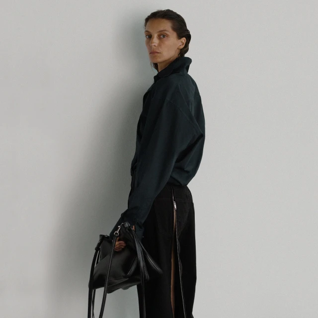 A woman in a black top and skirt holding a black bag stands in front of a white wall in a minimalist fashion shot.