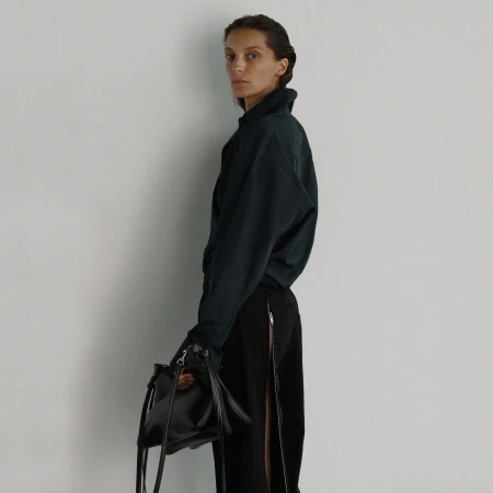 Model in dark button-up shirt and black trousers holding a black handbag against a light seamless background
