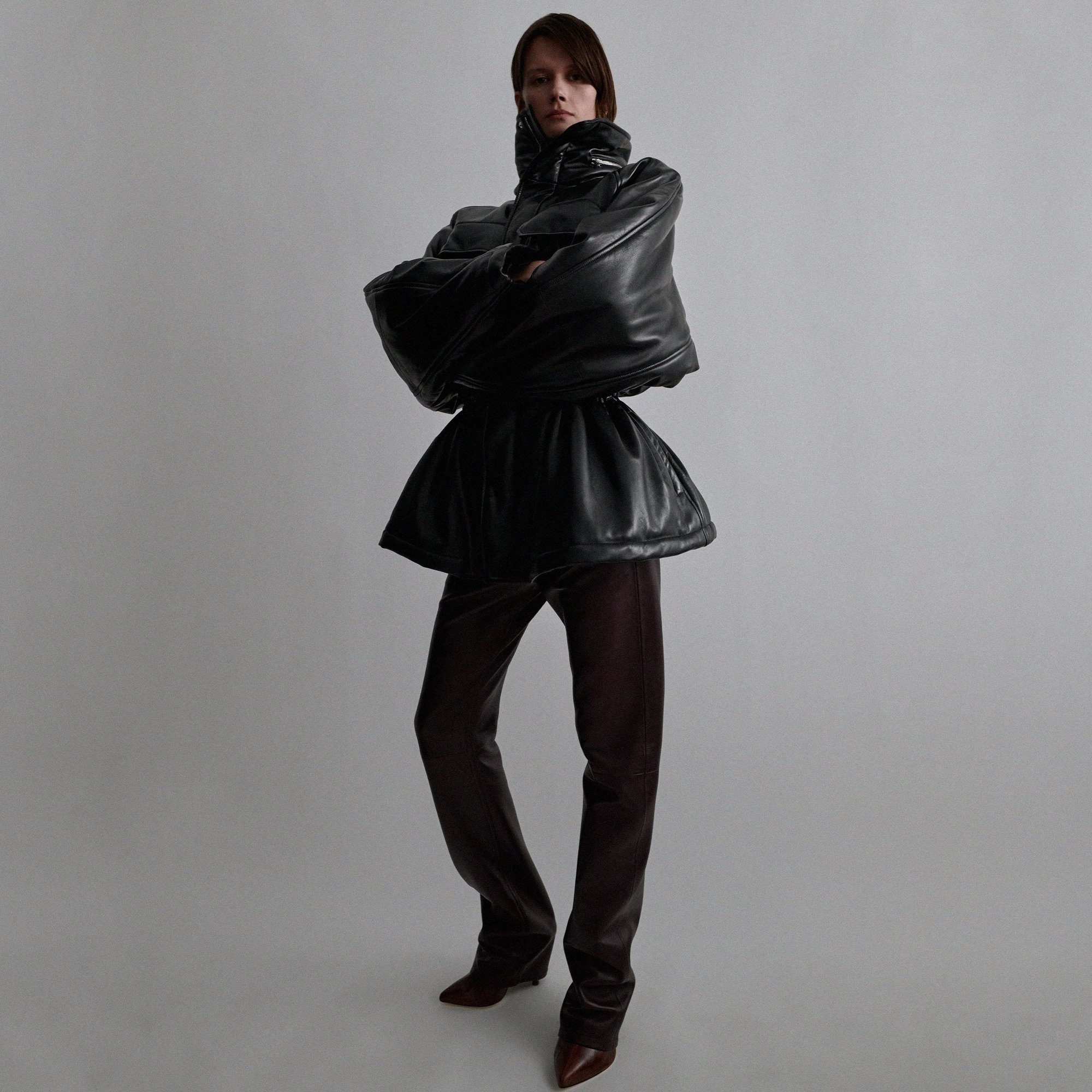 An adult woman poses in a high-fashion black leather jacket and maroon leather pants in a minimalist studio setting, full shot.