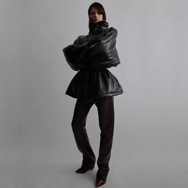 An adult woman poses in a high-fashion black leather jacket and maroon leather pants in a minimalist studio setting, full shot.