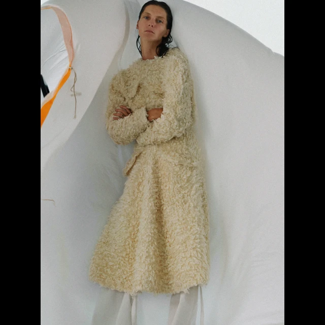 A woman in a shaggy coat stands in a brightly lit space with a white background, arms crossed, in a fashion editorial shot.