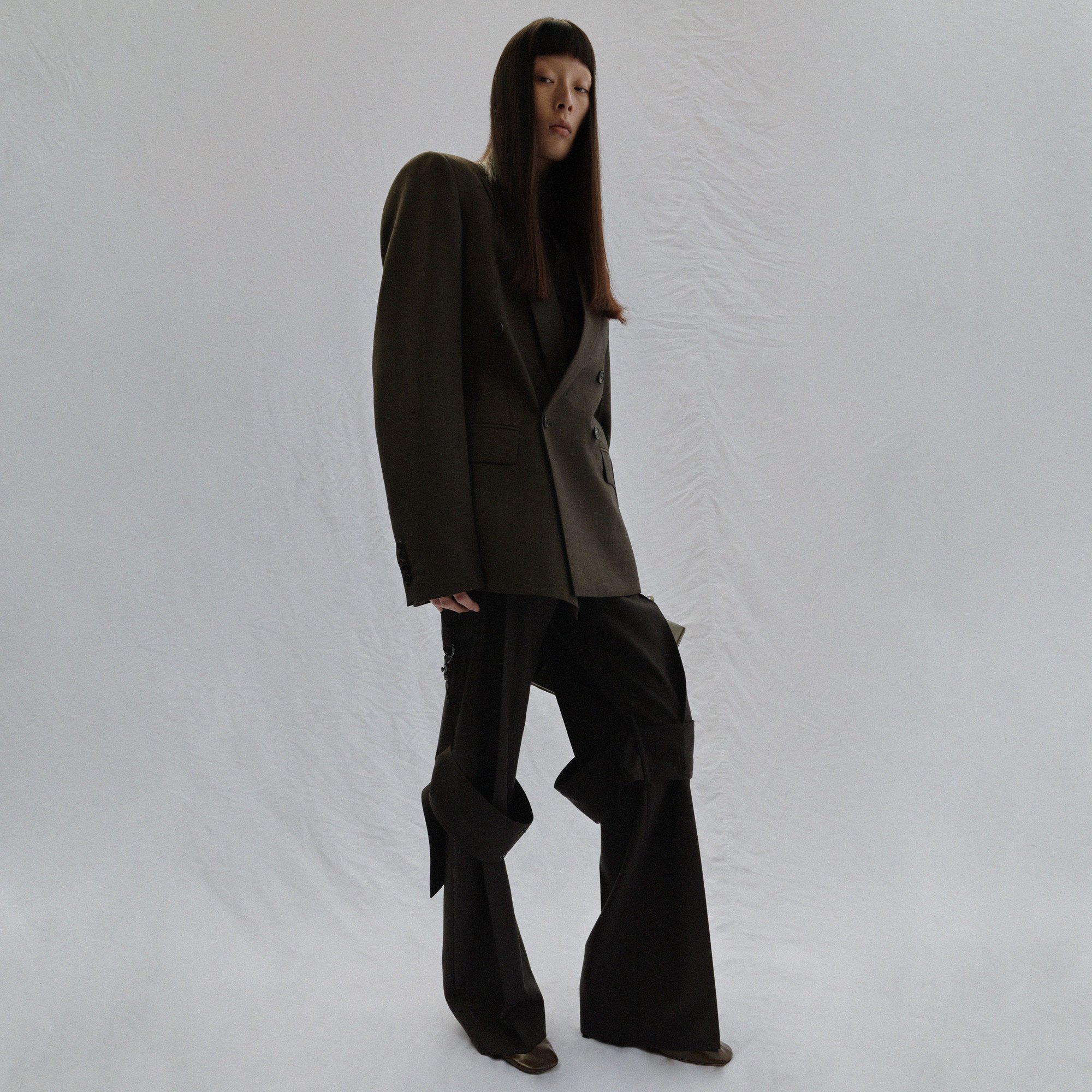 Asian woman stands in an olive-green double-breasted blazer and black pants with ties against a crinkled white backdrop in a studio shot.