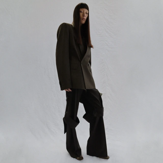 Asian woman stands in an olive-green double-breasted blazer and black pants with ties against a crinkled white backdrop in a studio shot.