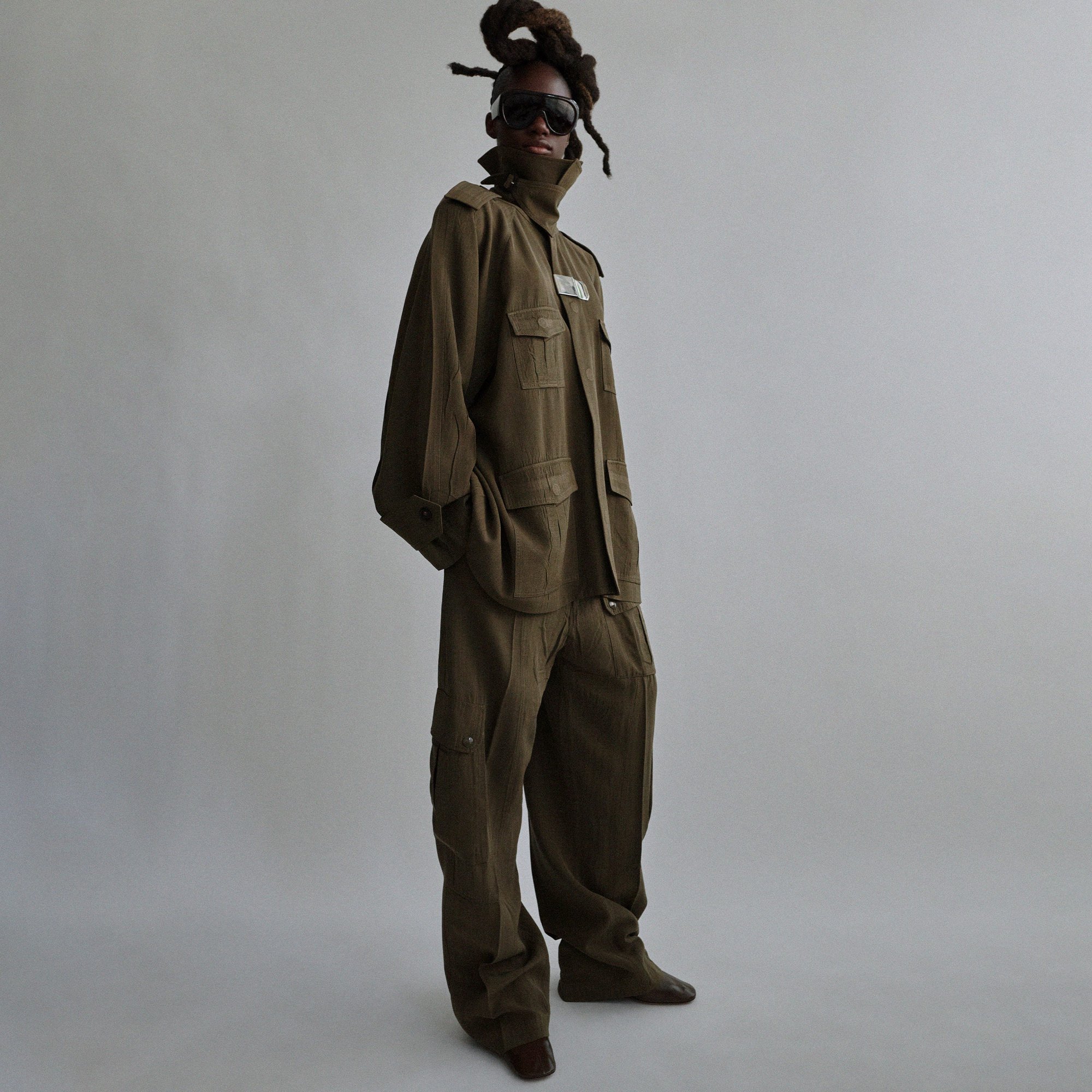 Full-length shot of an adult Black model in an olive-green outfit, sunglasses, and dreadlocks, standing in a studio with a neutral backdrop.