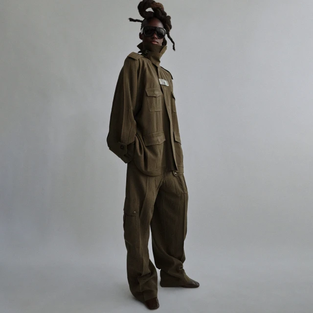 Full-length shot of an adult Black model in an olive-green outfit, sunglasses, and dreadlocks, standing in a studio with a neutral backdrop.
