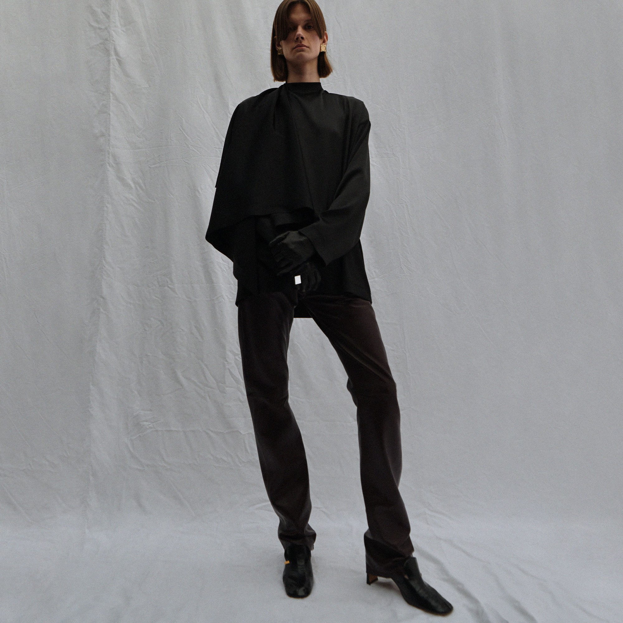 Full-length studio shot of a young adult posing in an oversized black top and brown pants against a white backdrop.