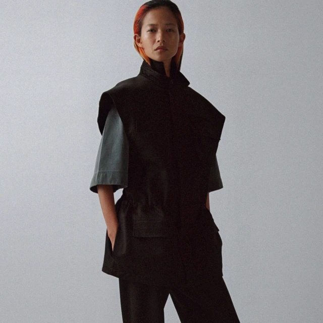 Fashion shot of a woman in a black vest and pants, with hands in pockets, set against a neutral backdrop in a studio setting.