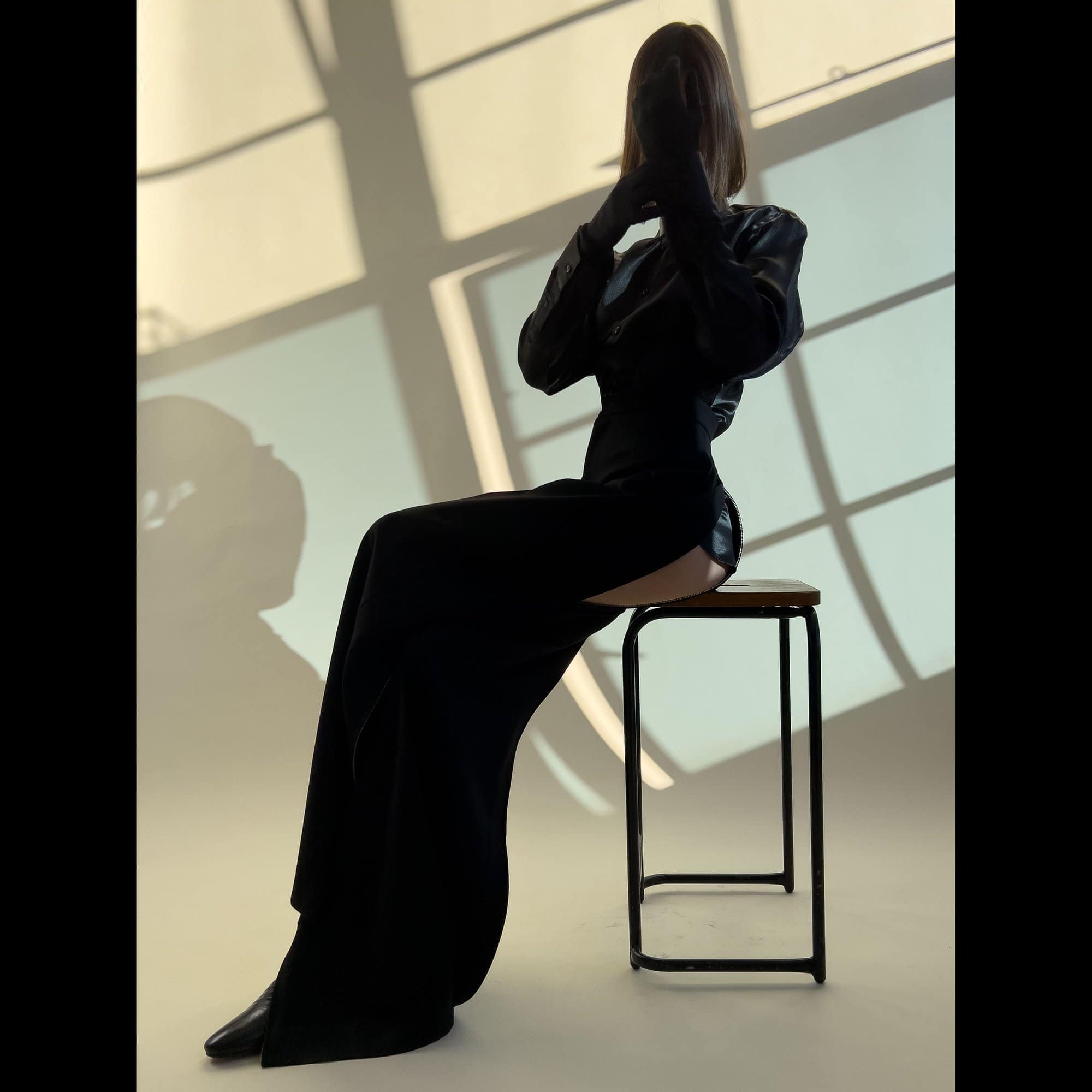 A woman in a black dress sitting on a stool in a studio with shadows, hands near her face, creating a silhouette, and stylish presentation.