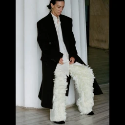 Model wearing a black coat and textured white feather trousers seated against large white columns.
