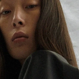 Close-up portrait of a model with long dark hair wearing a dark top against a neutral background.