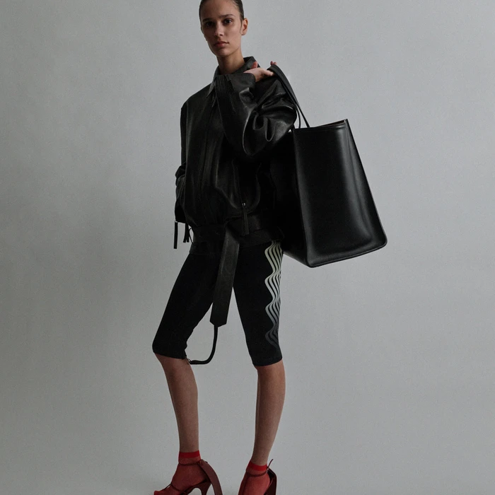 Model in a black leather jacket holding a large black tote bag against a neutral grey seamless studio background.