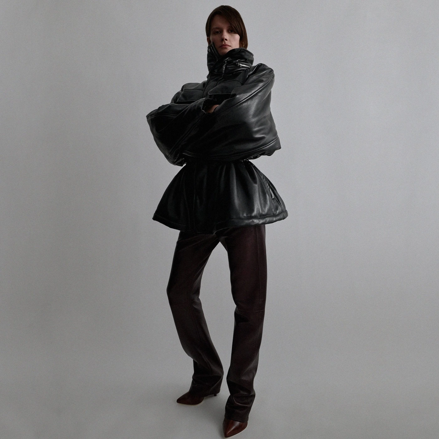 Model standing with arms crossed wearing a black leather puffer jacket and dark trousers against a grey seamless background